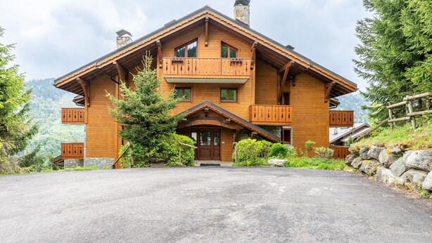 A large wooden chalet with a prominent balcony, a stone foundation, and a view of the surrounding forest.