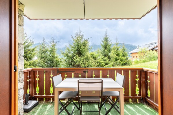 Une terrasse extérieure avec une table et des chaises, une clôture en bois agrémentée d’éléments décoratifs, et une vue sur les arbres et les montagnes.