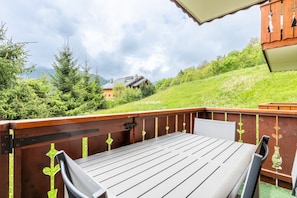 Une terrasse en bois avec une table et des chaises, entourée de verdure et avec un bâtiment visible au loin.