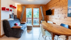 A cozy room with a sofa, ottoman, wooden dining table, chairs, a flat-screen TV, and a balcony with curtains.