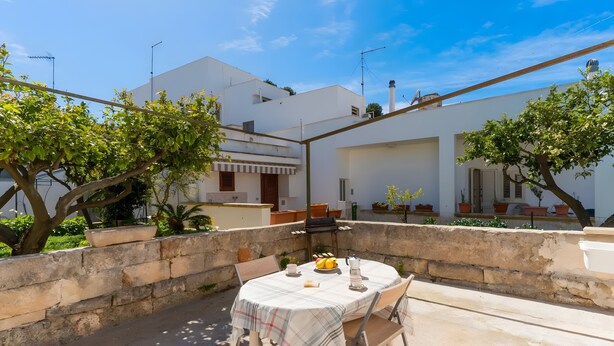 A patio with a table set for two, surrounded by a stone wall, with a white building in the background. - Cenate House Holiday Home (Nardò)