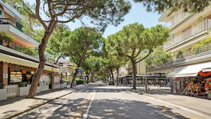A tree-lined street with buildings on either side, including shops and a café. - Apartment 'San Paolo 07' with Private Terrace and Air Conditioning (Lido di Jesolo)