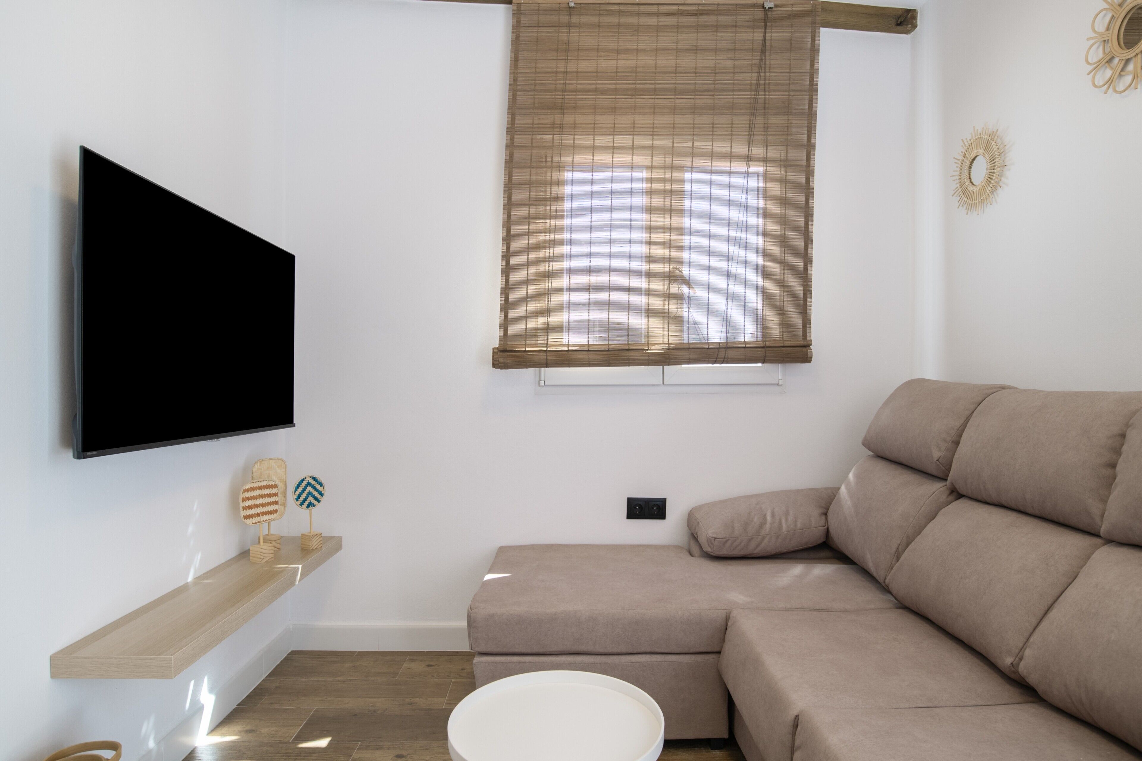 Un salon équipé d’un téléviseur à écran plat, d’un canapé beige, d’une étagère en bois et d’une fenêtre avec des stores.