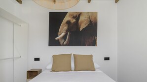 Un dormitorio con una cama, dos almohadas, un cuadro colgado en la pared de un elefante, una mesita de noche de madera y una lámpara en el techo.
