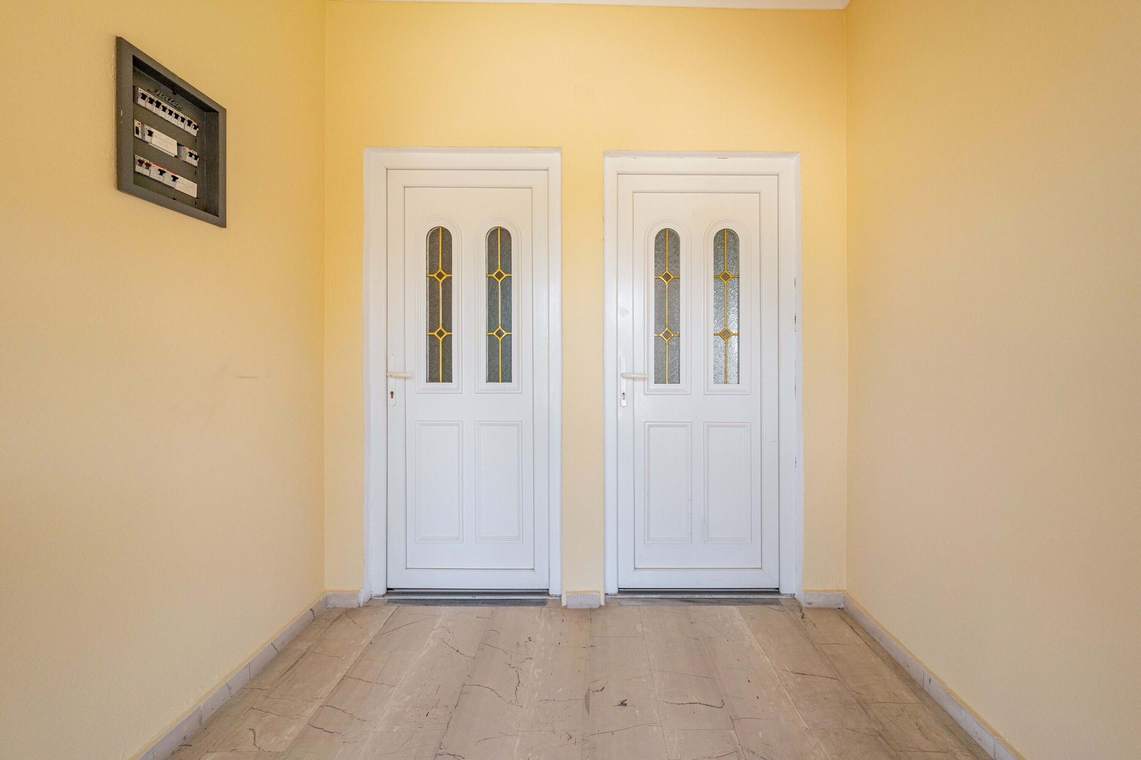 Un couloir avec deux portes blanches, un tableau électrique encadré et un sol carrelé.