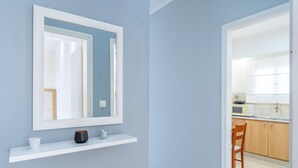 A room with a white-framed mirror, a white shelf with a white cup and a black mug, and a wooden chair in a kitchen area with wooden cabinets and a microwave.