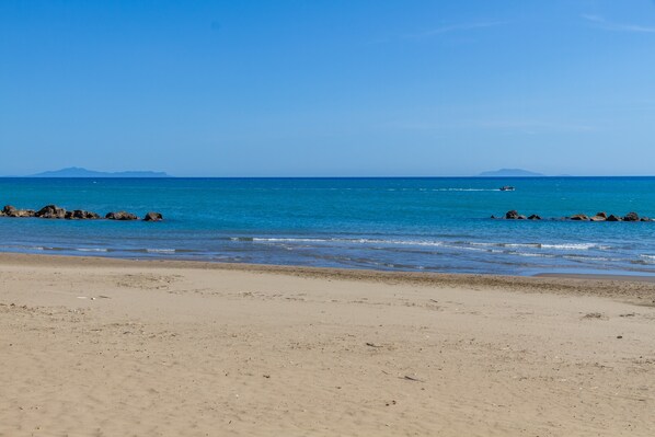 A beach with clear blue water, distant mountains, and a rocky shoreline.