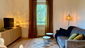 Sala de estar con televisión, lámpara, sofá y vista a la vegetación a través de una ventana.