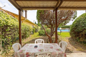 Un espacio de estar techado al aire libre con mesa y sillas, rodeado de vegetación y vistas a una piscina.