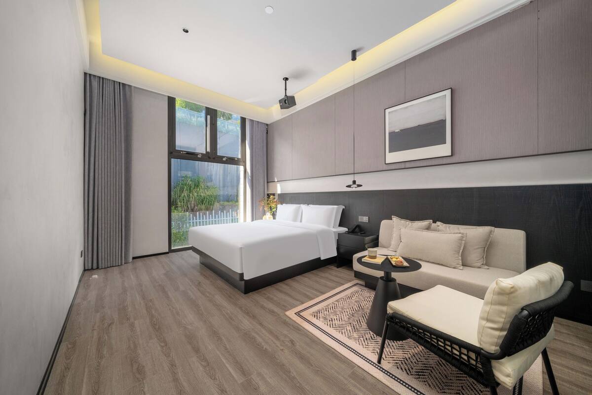 A modern hotel room with a large bed, a sofa, a small table, and a view of greenery outside.