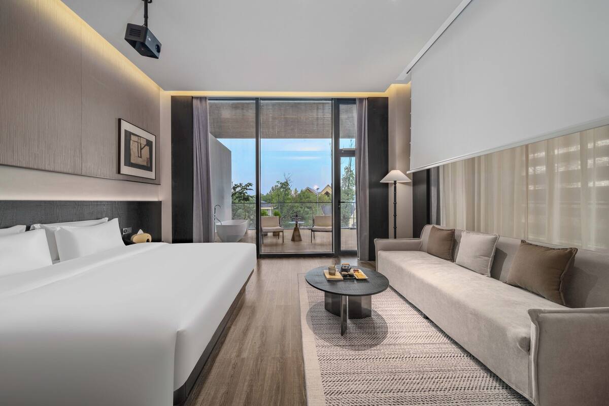 A modern hotel room with a large bed, a sofa, a coffee table, and a balcony with a view.