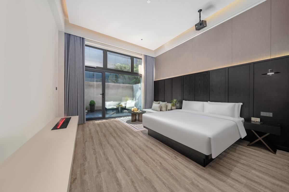 A modern hotel room with a large bed, a small desk, and a sliding glass door leading to a balcony.