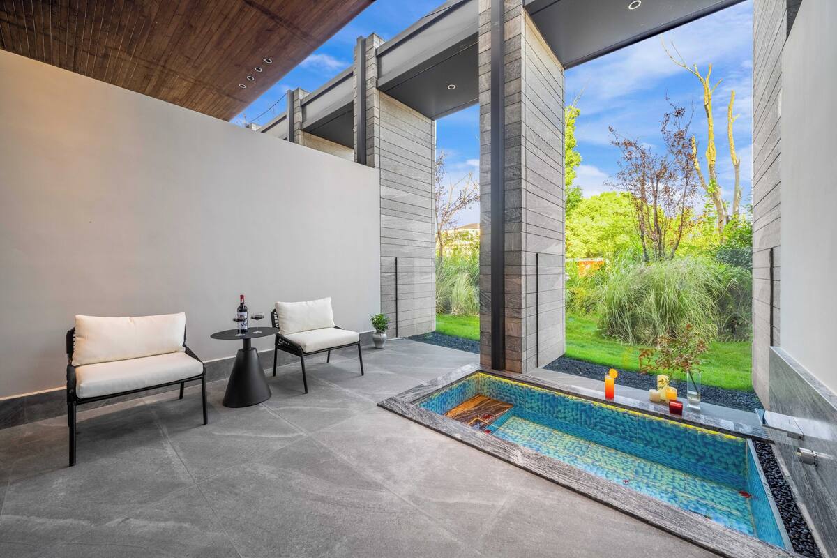 A modern outdoor seating area with a pool, two chairs, a table, and a view of a garden.