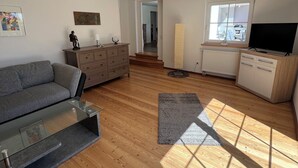 Amplia sala de estar con sofá, una mesita de centro de vidrio, televisor y una zona de cocina con armarios y ventana.