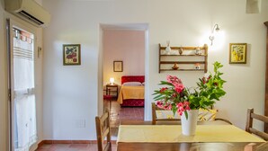 Un comedor con una mesa de madera, sillas, un florero con flores, un estante en la pared con adornos, y una cama roja al fondo.