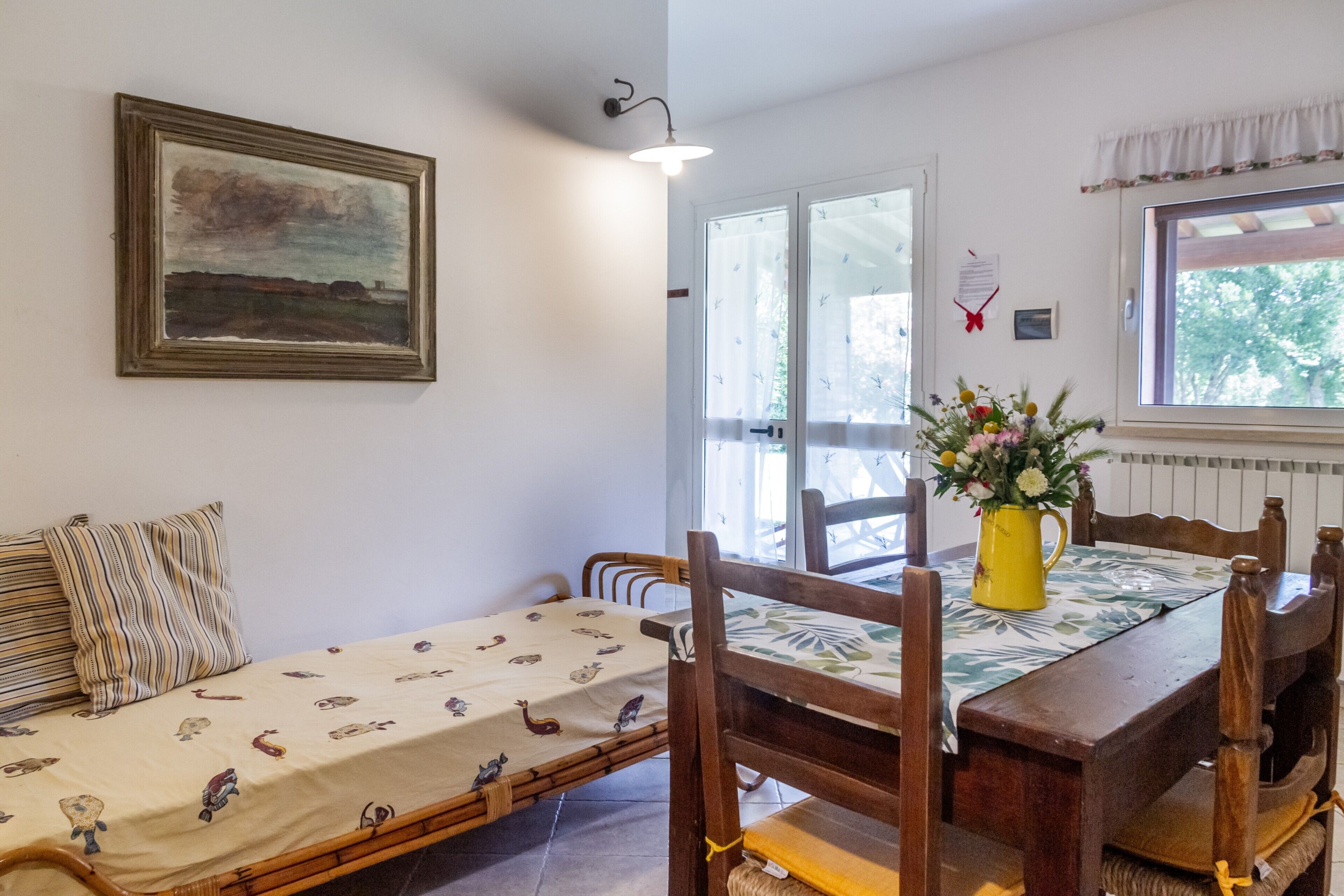 A room with a bed, a dining table with chairs, a vase with flowers, and a painting on the wall.