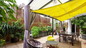 Een overdekt terras met een gele zonnescherm, een ronde tafel, stoelen, een fauteuil met kussens, potplanten en een houten omheining.
