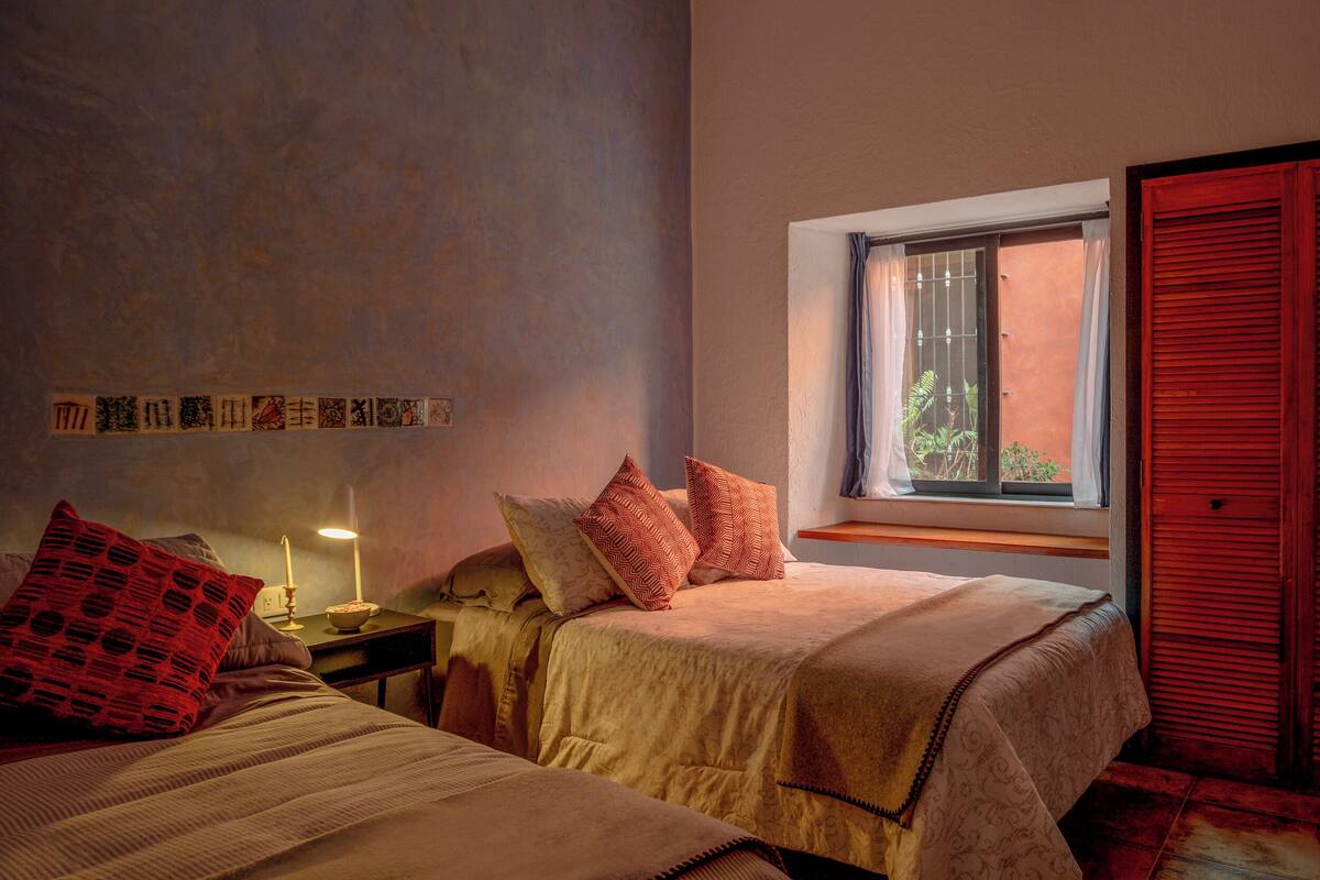 Ein Schlafzimmer mit zwei Betten, einem Nachttisch, einem Fenster mit Blick auf Grünflächen und einer roten Tür.