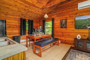 A wooden cabin interior with a kitchen, dining area, and a large window with a view of greenery. - Denton Go Cabin #3 | Denton Hill State Park (Ulysses)