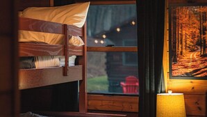 A cozy room with a bunk bed, a window with a view of a forest, and a lamp on a wooden table.