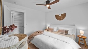 A bedroom with a bed, a ceiling fan, a wall-mounted decorative item, a bedside table with a lamp, and a door. - Wave Haven - Bali Style! Hot Tub! Near Bay & Beach (Kill Devil Hills)