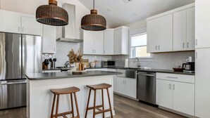 A modern kitchen with white cabinets, stainless steel appliances, and a central island with stools. - Wave Haven - Bali Style! Hot Tub! Near Bay & Beach (Kill Devil Hills)