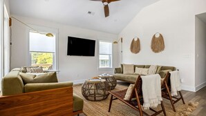 A modern living room with a sofa, armchair, and coffee table, a flat-screen TV mounted on the wall, and a ceiling fan. - Wave Haven - Bali Style! Hot Tub! Near Bay & Beach (Kill Devil Hills)