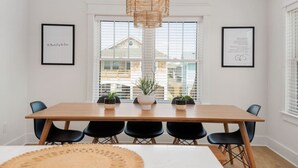 Un coin repas avec une longue table en bois, des chaises noires et un luminaire suspendu en fibres tressées. Les murs sont ornés de tableaux encadrés et la table est décorée de plantes en pot.