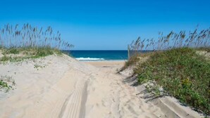 Un sentier de sable qui longe la plage et mène à l’océan, bordé de dunes herbeuses.