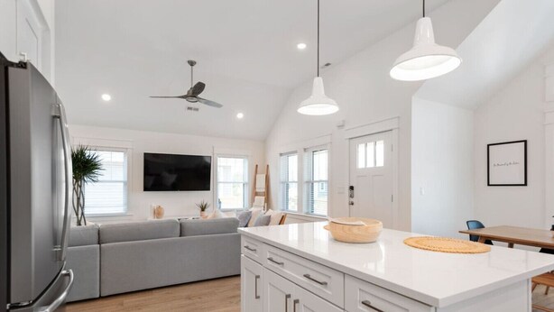 Une cuisine moderne avec des meubles blancs, un canapé gris, un coin repas avec une table en bois et un ventilateur de plafond.