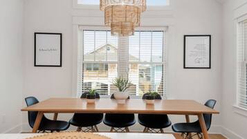 A dining area with a long wooden table, black chairs, and a woven pendant light. There are framed pictures on the walls and potted plants on the table.