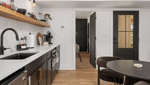A modern kitchen with a black faucet, white countertop, and wooden shelves. There is a dining area with a round table and chairs. - Cocoa Capri - Luxury Getaway (Kill Devil Hills)