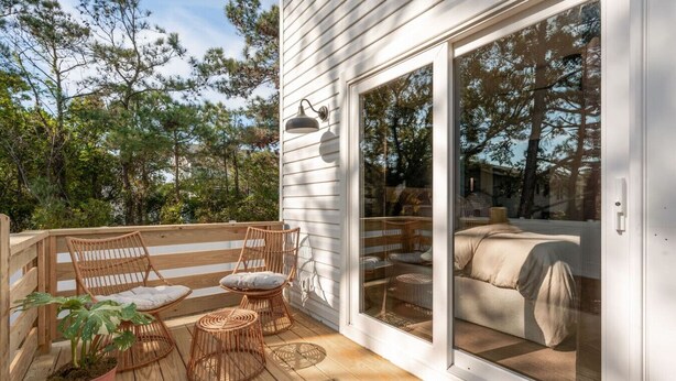 A wooden deck with wicker furniture, a potted plant, and a sliding glass door leading to a bedroom with a bed and a chair. - Cocoa Capri - Luxury Getaway (Kill Devil Hills)