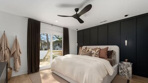 A bedroom with a bed, a ceiling fan, a nightstand, and a view through a sliding door. - Cocoa Capri - Luxury Getaway (Kill Devil Hills)