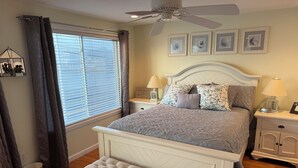 Un dormitorio con cama, mesitas de noche, un banco, ventilador de techo y ventana con persianas.