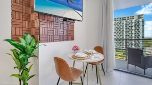 A dining area with a TV mounted on a brick wall, a balcony with a view of a high-rise building, and a potted plant. - Stylish Studio | Pool View | Downtown Doral' Heart (Doral)