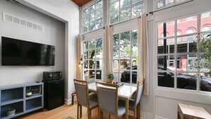 Una sala de estar moderna con un televisor, una mesa de comedor con sillas, un mueble negro pequeño y un ventanal grande con cortinas.