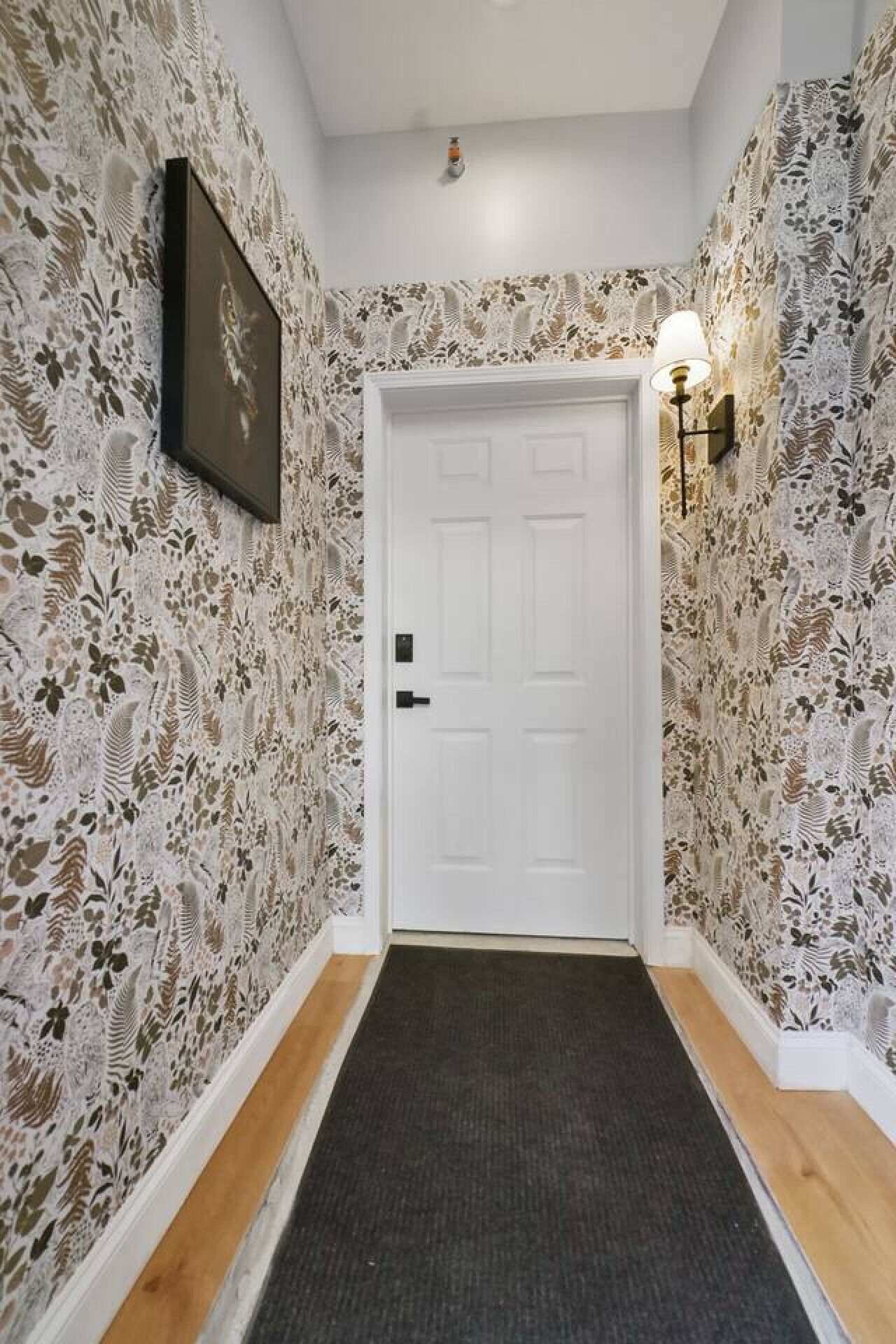 A hallway with patterned wallpaper, a white door, a framed picture on the wall, and a wall-mounted light fixture.