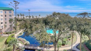 A resort with a pool surrounded by trees, a beach view, and a building with balconies.