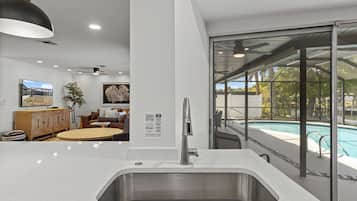 A modern kitchen with a stainless steel sink, a dining area with a round table, and a view of a swimming pool through large glass doors.
