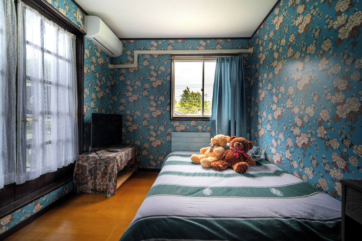 A bedroom with floral wallpaper, a bed with a striped bedspread, a window with curtains, and a television on a small table.