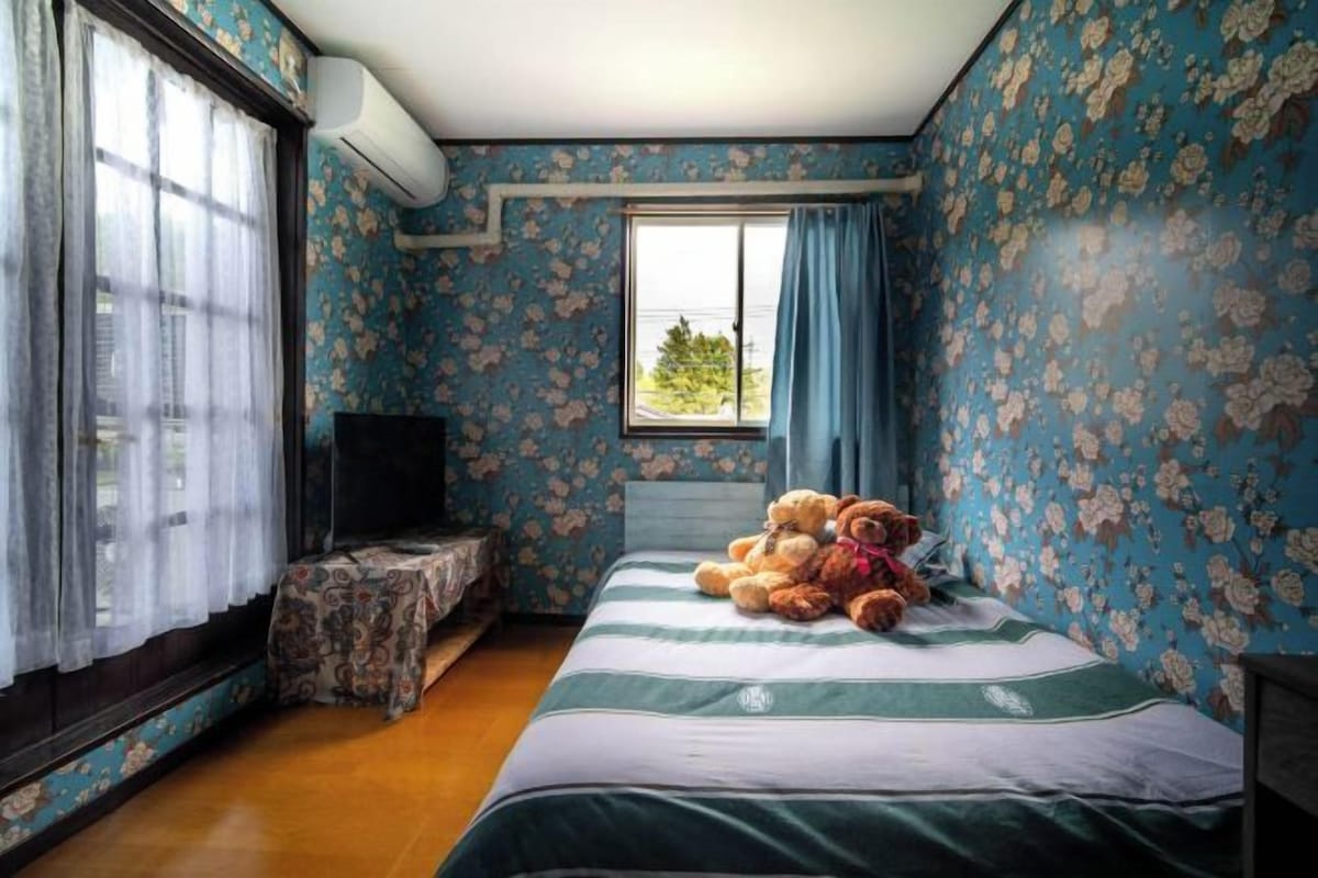 A bedroom with floral wallpaper, a bed with a striped bedspread, a window with curtains, and a television on a stand.