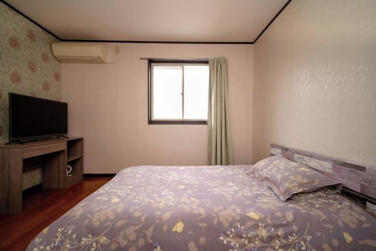 A bedroom with a bed, a television on a stand, a window with curtains, and a wall-mounted air conditioner.