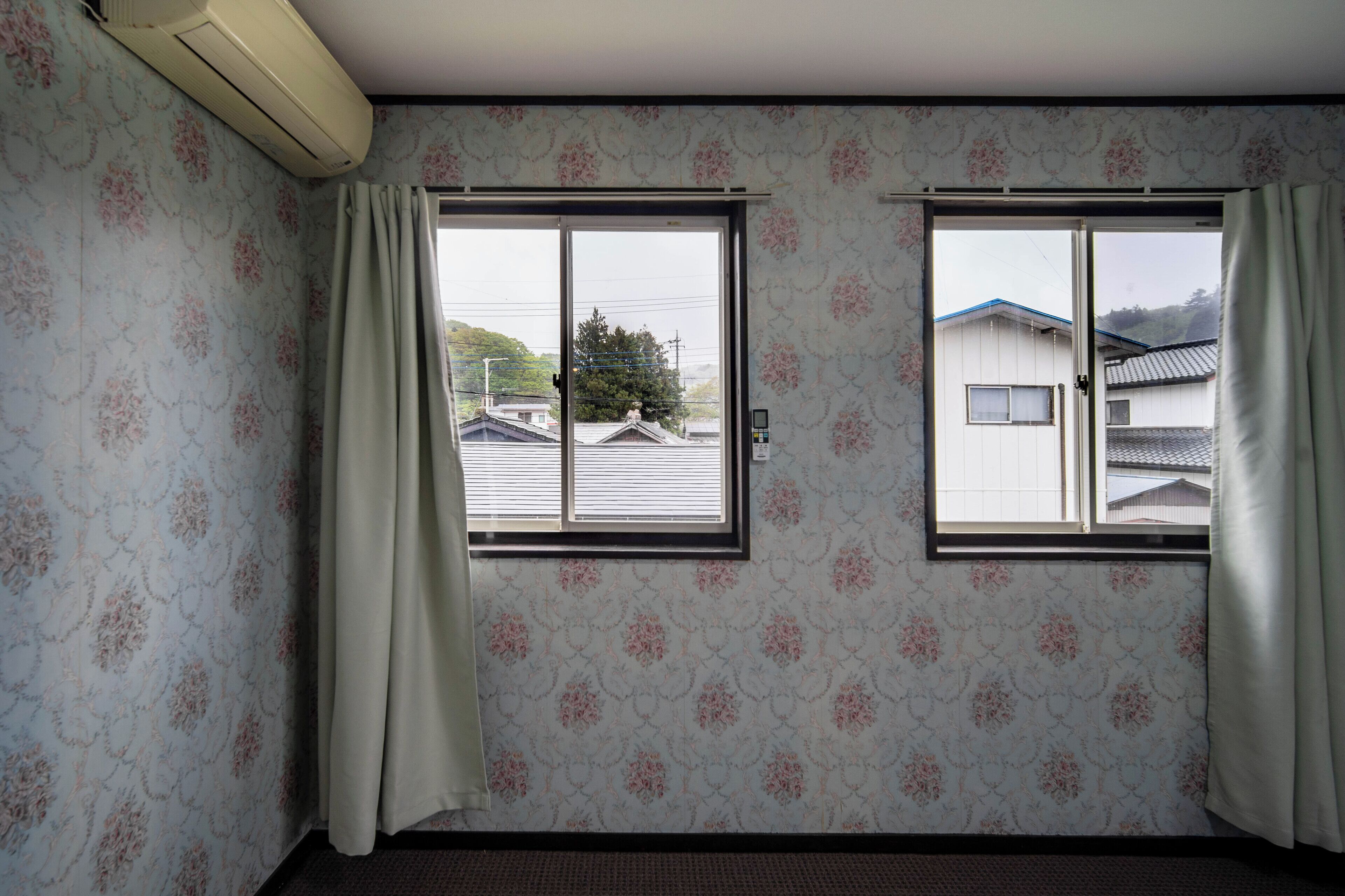 A room with floral wallpaper, two windows with curtains, and a view of houses outside.