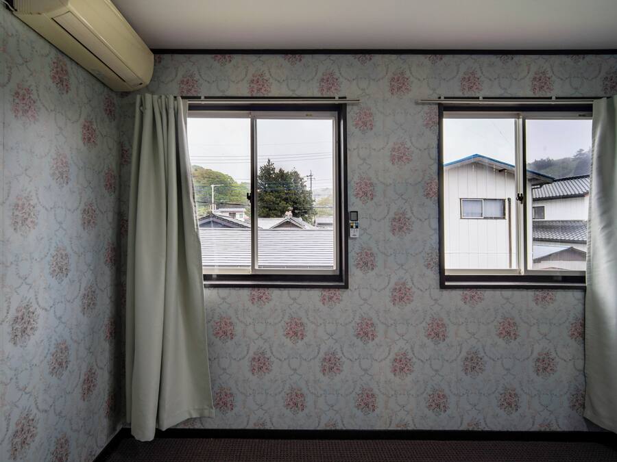 A room with floral wallpaper, two windows with curtains, and a view of houses outside.