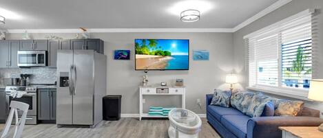A modern living room with a blue sofa, a flat-screen TV, and a kitchen area with stainless steel appliances.