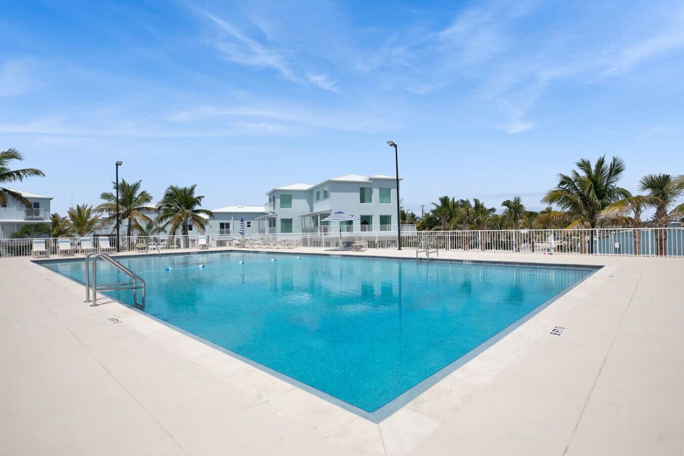 A rectangular swimming pool with a metal ladder on one side, surrounded by a white fence. There are palm trees and a building with balconies in the background.