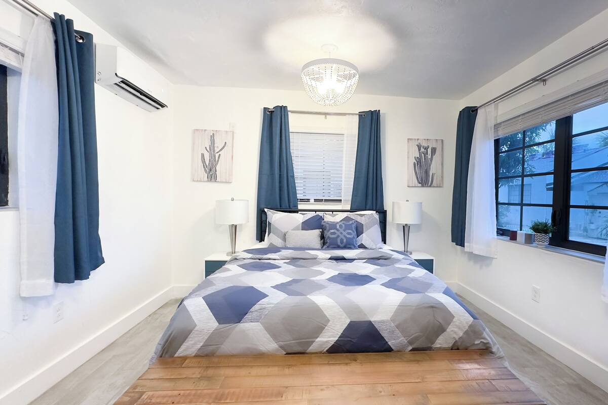 A bedroom with a bed, two bedside lamps, a chandelier, and a window with curtains.