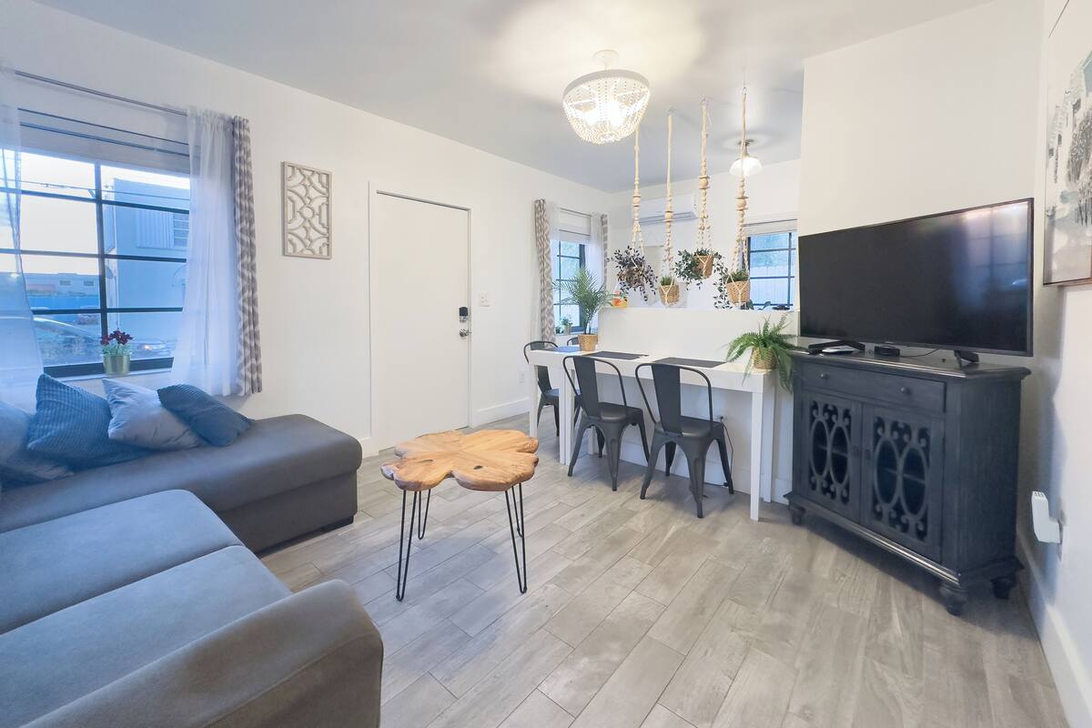 A modern living room with a sofa, a coffee table, and a TV. There is a dining area with chairs and a table, and a kitchen area with a TV and plants.