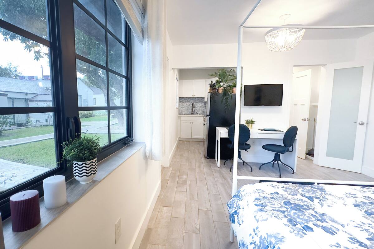 A modern bedroom with a large window, a bed, a desk, and a TV.